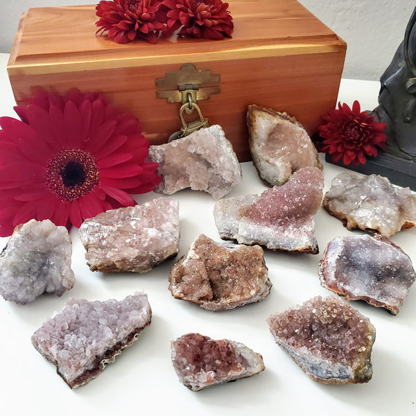 Pink Amethyst Clusters in a variety of sizes in front of a wooden box and Buddha statue, with accents of fresh red flowers