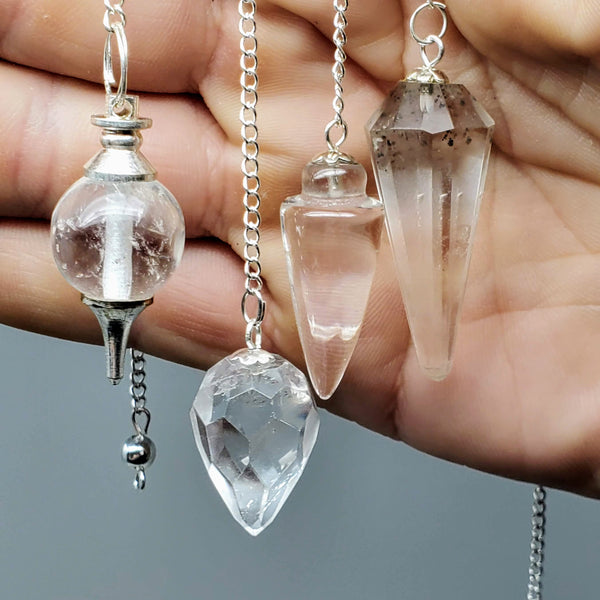 Close-up of 4 different styles of Clear Quartz pendulums draped over a hand on a gray background
