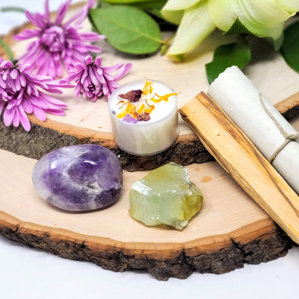 The contents of our Let That Sh*t Go Evening Meditation Ritual, including a Chevron Amethyst palm stone, a raw Green Calcite chunk, a crystal infused tea light candle, smudge stick, and guided meditation scroll, displayed on natural wood slabs with fresh flowers in the background