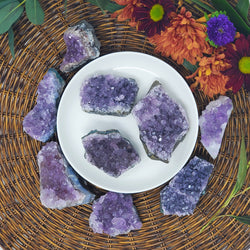 A variety of sizes of Amethyst Clusters on and around a white tray, with a brown wicker charger underneath and fresh flowers behind them