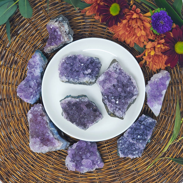 A variety of sizes of Amethyst Clusters on and around a white tray, with a brown wicker charger underneath and fresh flowers behind them