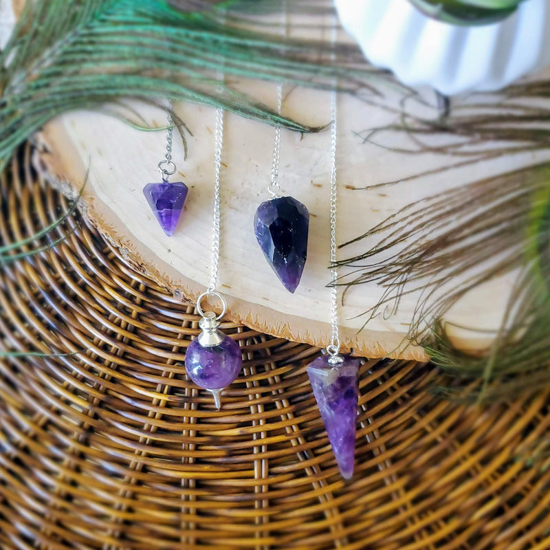 4 different styles of Amethyst pendulums gently draped over a natural wood slab on a brown wicker background, all framed by peacock feathers.