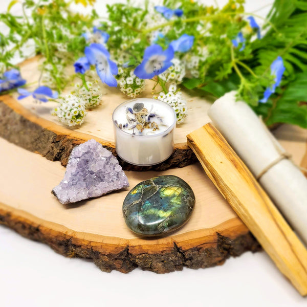 Chill The F*ck Out Evening Meditation Ritual kit includes Lavender Amethyst, Labradorite palmstone, 8-hour tea light candle, smudge stick, and meditation script as shown here, laid out on stacked natural wood slabs with flowers in the background
