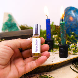 A hand holding up a bottle of Balance Solution Meditation Oil in front of two burning candles that are dressed with herbs, as well as a Fluorite slab, a Blue Apatite freeform, and flowers 