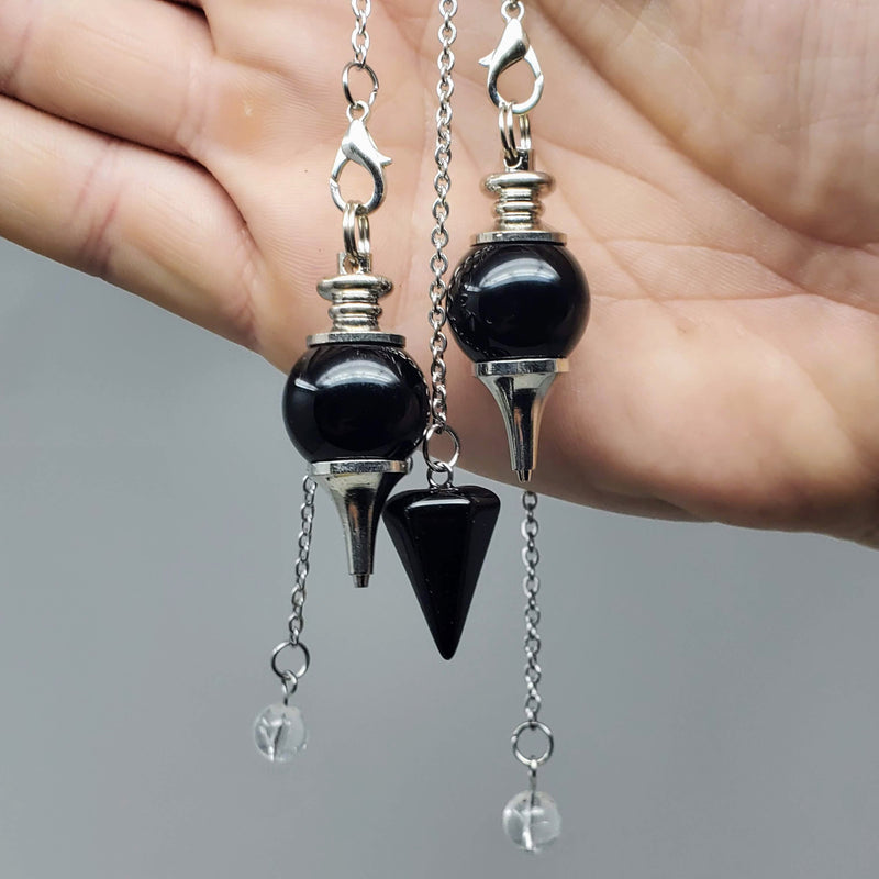 2 round and 1 triangle style Black Obsidian pendulums draped over an open palm on a gray background