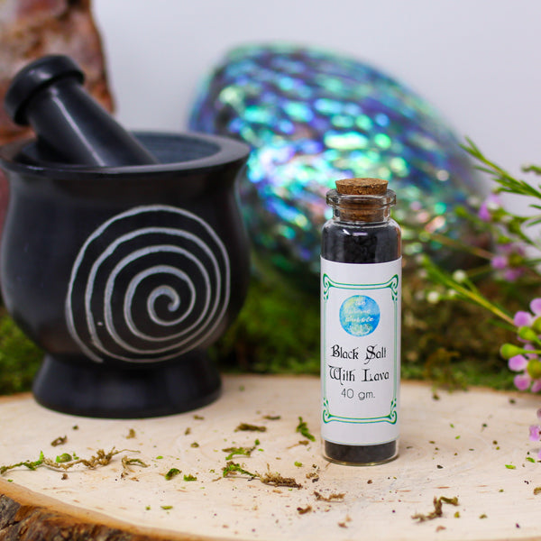 A bottle of Black Salt with Lava with a magical background that includes an abalone shell, Pink Amethyst, mortar and pestle, and fresh flowers 