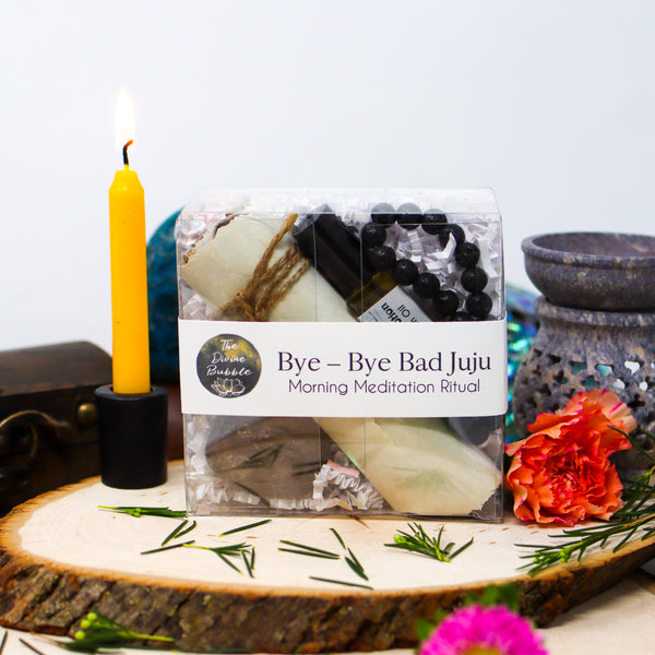 Our Bye-Bye Bad Juju Morning Meditation Ritual kit nestled between a lit yellow candle and an oil burner, all sitting on a natural wood slab in front of a white background