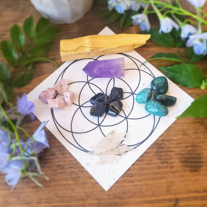 Call in calm Ritual Grid Kit includes Amethyst generator, Quartz points, palo santo, Rhodonite, Chrysocolla, Tiger's Eye, and a crystal grid that is printed on seed paper, all seen here laid out on a stained wood slab surrounded by fresh flowers