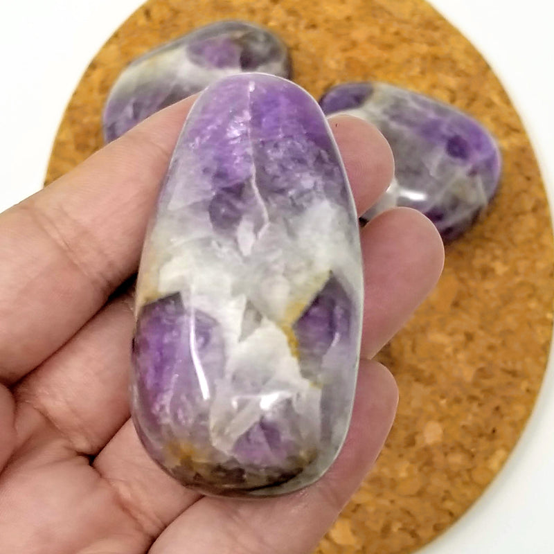 A close up view of a Chevron Amethyst palmstone resting on a hand, with more palmstones on a cork backdrop
