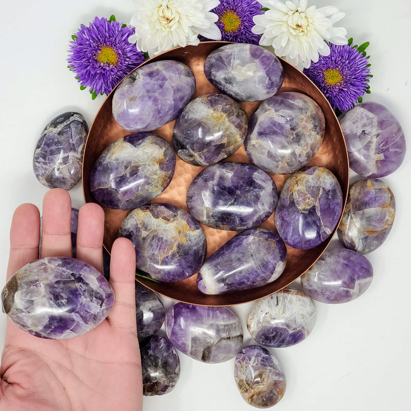 An open hand holding a Chevron Amethyst palmstone, with a variety of sizes of Chevron Amethyst palmstones in and around a hammered copper bowl in the background, accented with purple and white flowers