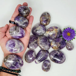 5 different sizes of Chevron Amethyst palmstones, arranged from smallest to largest, laid on an open hand, with more varied sizes of palmstones in the background