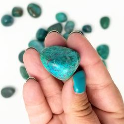 A close up of a Chrysocolla tumbled stone held in a hand, with more tumbled Chrysocolla in the background