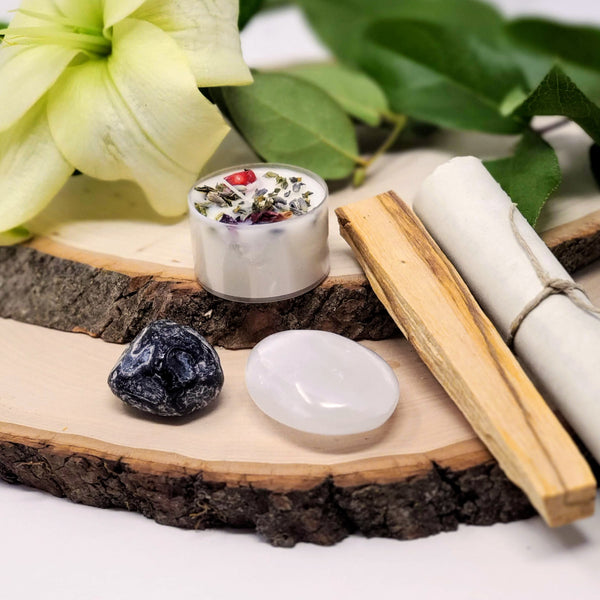 The contents of our My Safe Space Evening Meditation Ritual, including 8 hour meditation candle, palo santo, meditation scroll, Selenite palm stone and Apache tear displayed on natural wood slabs with fresh flowers in the background 