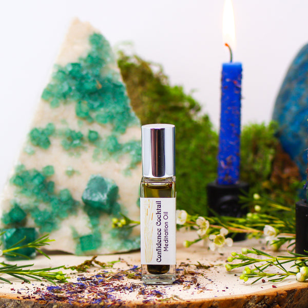 A bottle of our Confidence Cocktail Meditation Oil standing in front of a slab of Fluorite and a burning blue candle dressed with herbs.