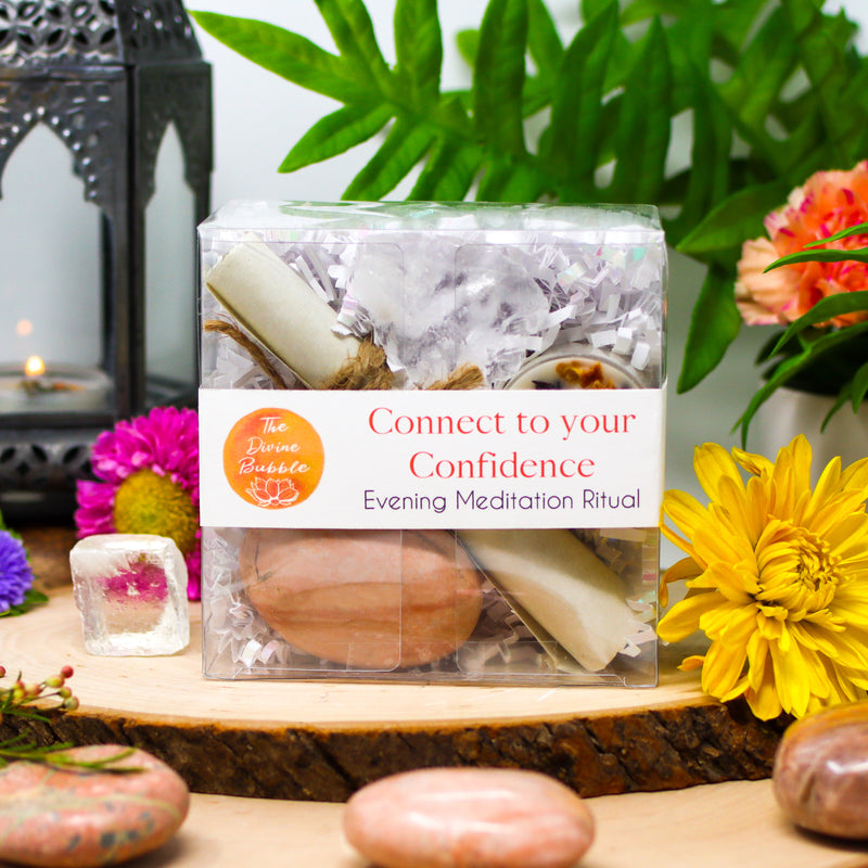 Our Connect To Your Confidence Evening Meditation Ritual kit surrounded by beautiful fresh flowers, Peach Moonstone palm stones, and a raw White Calcite chunk, with a burning lantern in the background