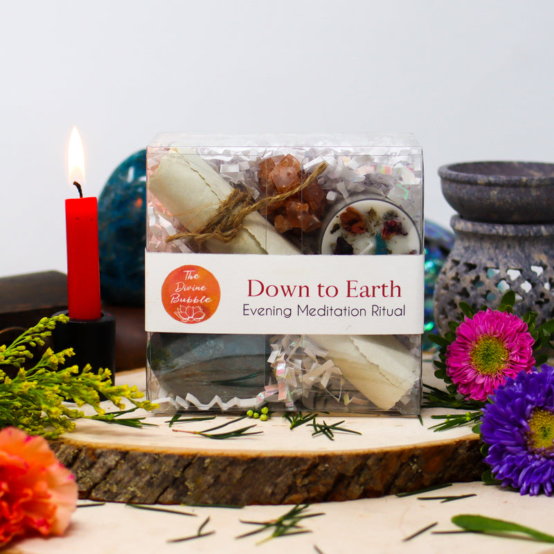 Our Down to Earth Evening Meditation Ritual kit, lovingly displayed on two stacked natural wood slabs that are surrounded by vibrant fresh flowers and greenery, a burning red candle, Blue Apatite freeform, and an oil burner