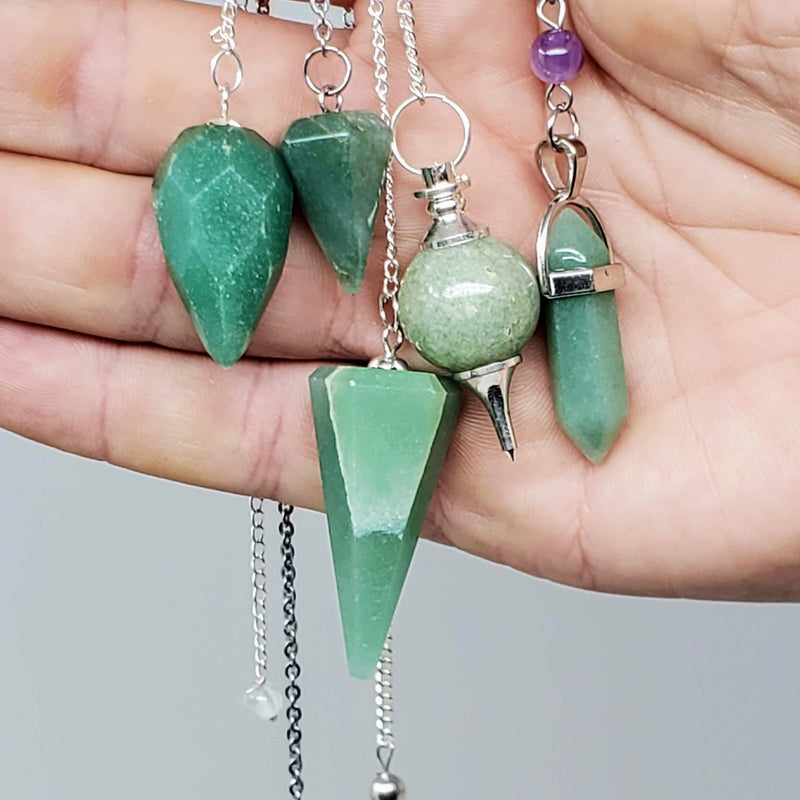Close-up of 5 different styles of Green Aventurine pendulums draped over the palm of a hand on a gray background