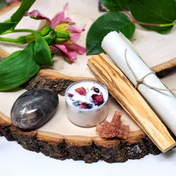 Our Down to Earth Evening Meditation Ritual includes Black Moonstone palm stone, Aragonite star, Meditation Scroll, 8-hour meditation candle, and smudge stick, as seen here on two stacked natural wood slabs with fresh flowers in the background 