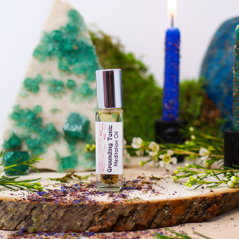 A bottle of our Grounding Tonic Anointing Oil in front a slab of Fluorite and a burning candle dressed in herbs, all atop a natural wood slab