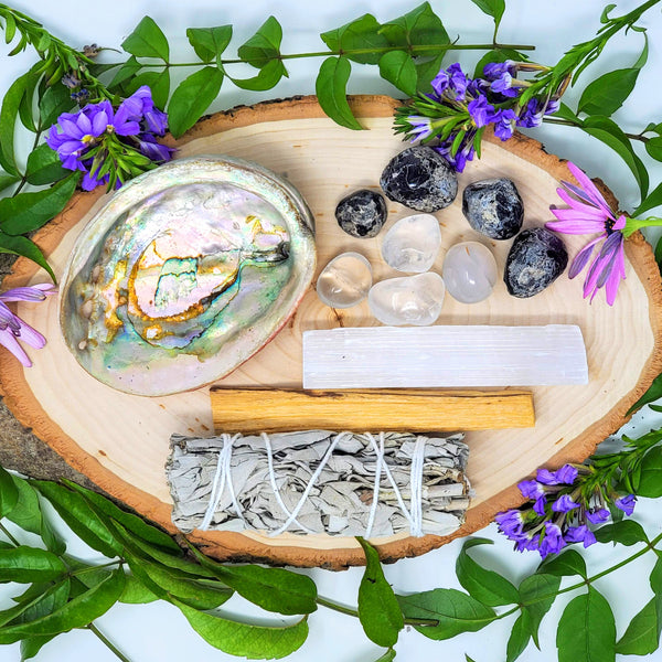 The contents of our Bless your Space Crystal Smudge Ritual, including white sage, palo santo, 4 Apache tears, 4 Clear Quartz, selenite stick, and abalone shell beautifully laid out on a natural wood slab surrounded by flowers