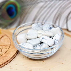 A clear glass bowl filled to the brim with Howlite Tumbled Stones on a natural wood slab with a peacock feather in the background