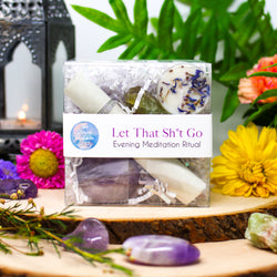 Our Let That Sh*t Go Evening Meditation Ritual displayed on two stacked natural wood slabs and surrounded by Chevron Amethysts, raw Green Calcite chunks, and beautiful fresh flowers, with a lantern burning in the background