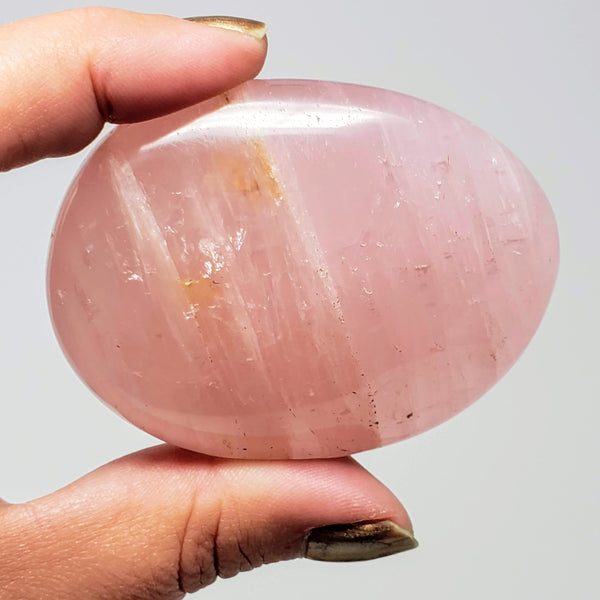 A close up of a hand holding up a Rose Quartz palmstone with a white background