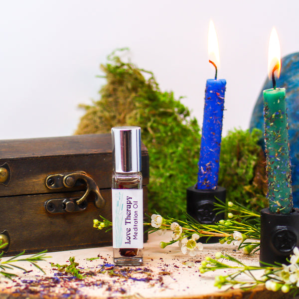 A bottle of Love Therapy Anointing Oil in front of a wooden box and two burning candles dressed in herbs