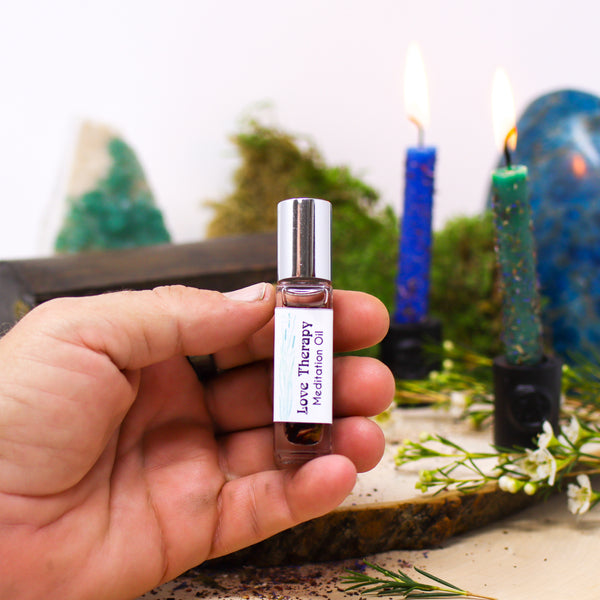 A hand holding a bottle of Love Therapy Anointing Oil with two dressed candles and a Blue Apatite freeform in the background