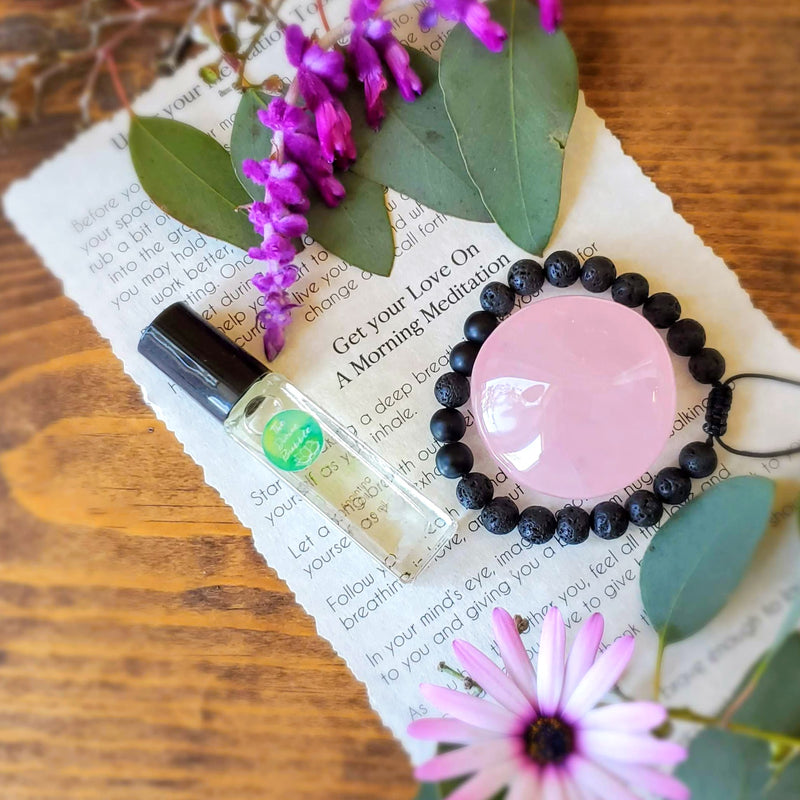 Lovely display of Get your Love On Morning Meditation Ritual Kit contents, including Rose Quartz palmstone, diffuser bracelet, anointing oil, and guided meditation laid out on a stained wood background, framed by stunning fresh flowers