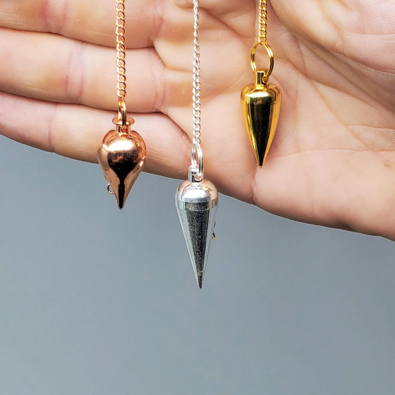 One of each variety (copper, silver, and brass) of metal pendulums draped over the side of a hand on a white background