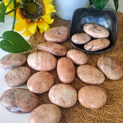 Various sizes of Peach Moonstone Palm Stones spilling out of a square bowl onto a brown scalloped straw backdrop, with sunflowers in the background 
