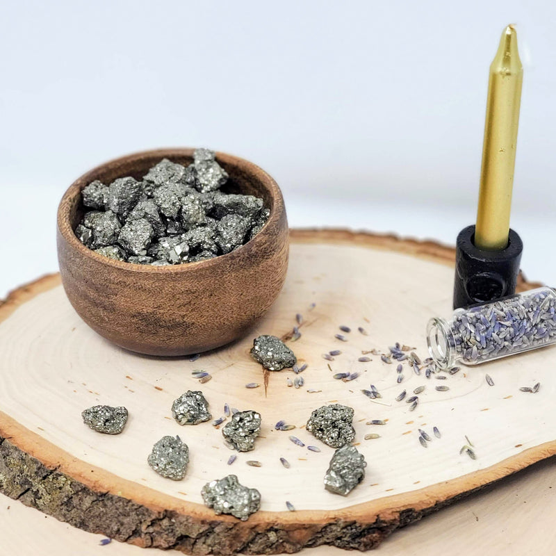 A gorgeous display of Pyrite Baby Clusters, some in a wooden bowl and others scattered about on top of a natural wood slab, along with an open bottle of dried lavender that has been sprinkled out, as well as a metallic gold chime candle in a cast iron candle holder, all on a white background