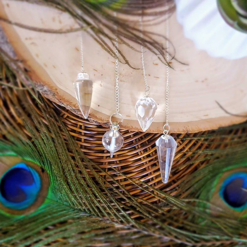 4 different styles of Clear Quartz pendulums draped over the side of a natural wood slab that is set on a brown wicker charger, surrounded by peacock feathers 