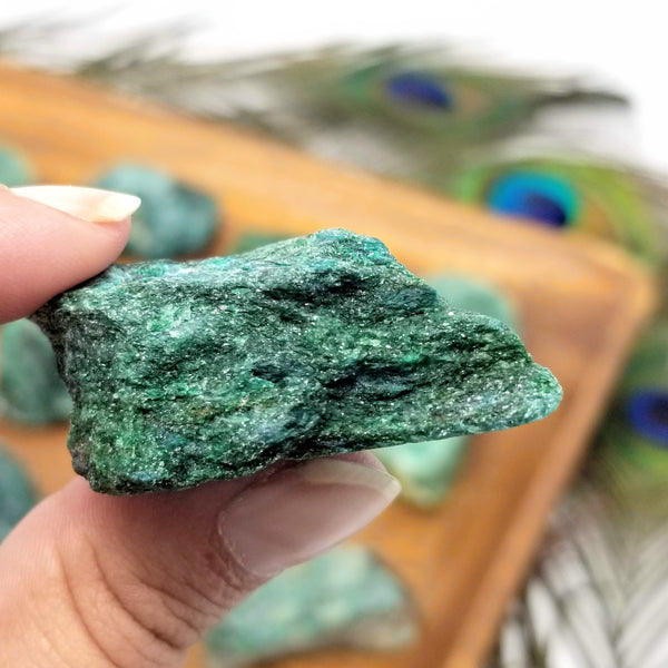 A sparkly Raw Fuchsite held up between a forefinger and thumb in front of a wooden tray full of Fuchsite, with peacock feathers in the background