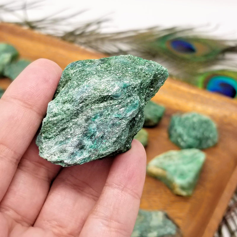 A sparkly Raw Fuchsite resting on the fingertips of an open hand in front of a wooden tray full of Fuchsite, with peacock feathers in the background