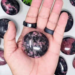 An open hand holding a Rhodonite palmstone, with a stunning array of Rhodonite palm stones behind it, on a white background