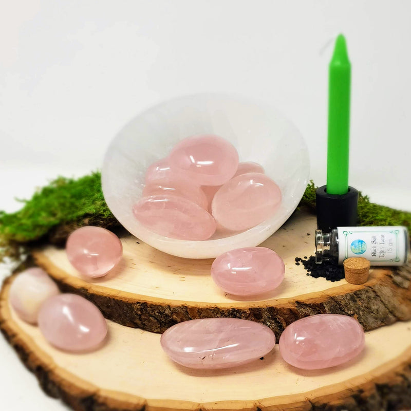Rose Quartz palmstones displayed in a Selenite bowl set atop two natural wood slabs, with additional Rose Quartz palmstones in the foreground, an uncorked bottle of Black Salt with Lava on its side with the contents spilling out and a green chime candle in a cast iron candle holder to the side, all with a white background