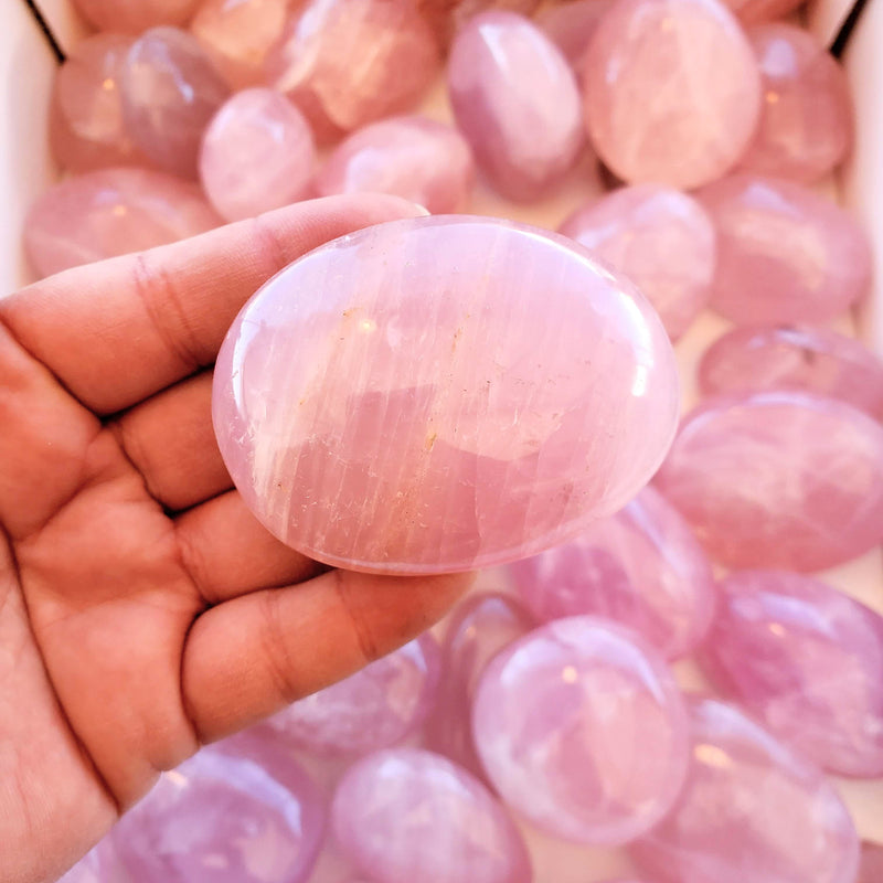 A close up of an open hand holding a Rose Quartz palmstone, with an abundance of the most stunning Rose Quartz palmstones in the background