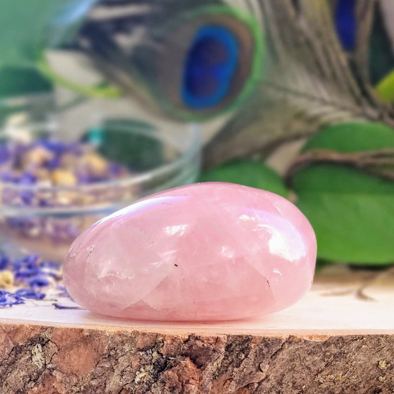 A close up of a beautiful Rose Quartz palmstone, with a clear bowl of dried Corn Flowers spilling over and peacock feathers in the background