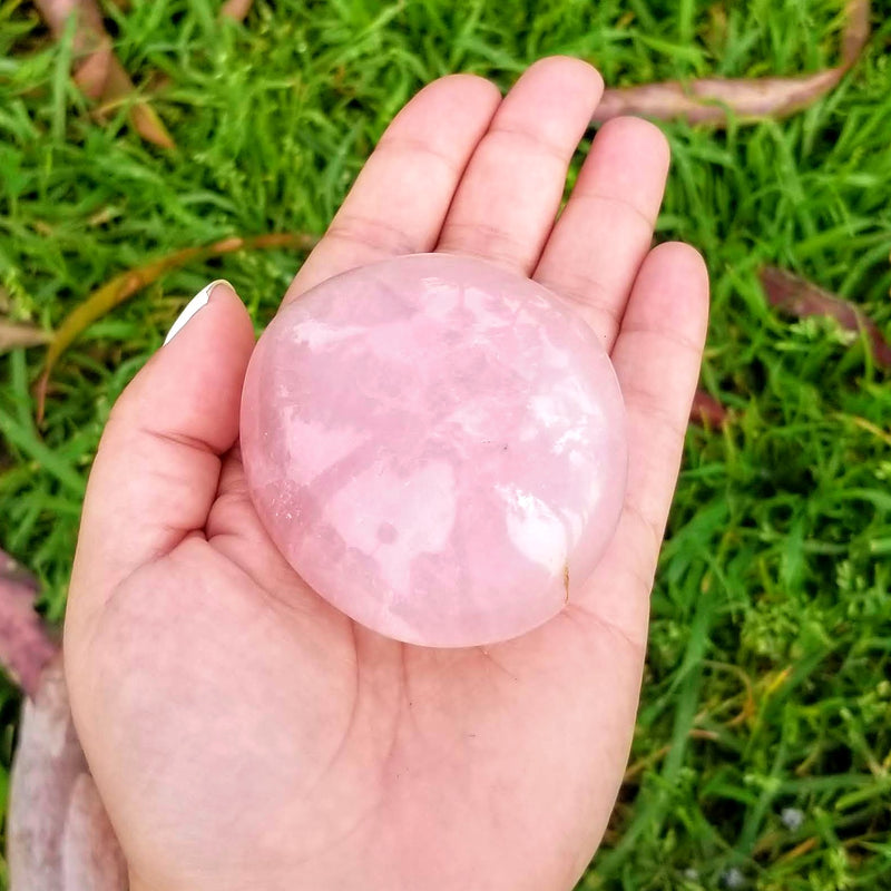 An open hand with a Rose Quartz palmstone lying in the palm, with grass and leaves in the background