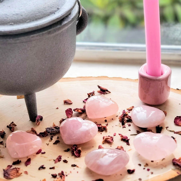 Rose Quartz Pocket Hearts scattered across a natural wood slab along with dried Red Rose petals, as well as a cast iron cauldron and a pink chime candle in a pink candle holder