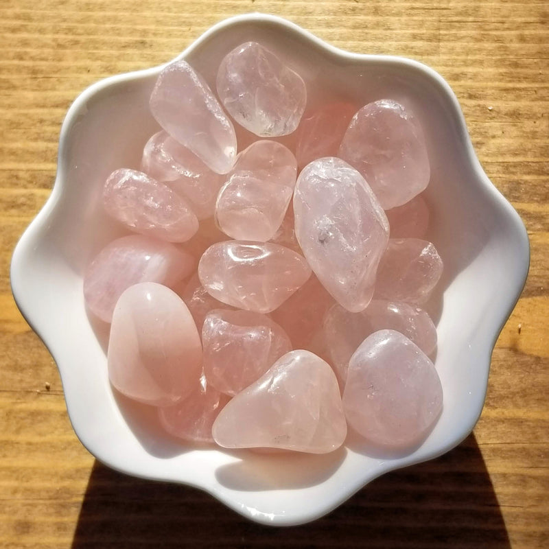 A white bowl with scalloped edges filled with Rose Quartz tumbled stones in a variety of sizes on a stained wood background 