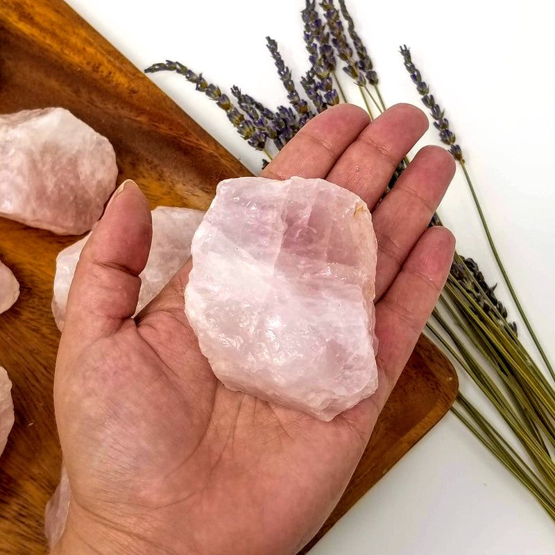 A Rose Quartz Raw Chunk resting in the palm of an open hand, with more Rose Quartz Raw Chunks in a variety of sizes on a stained wood tray with purple flowers to the side, on a white background 
