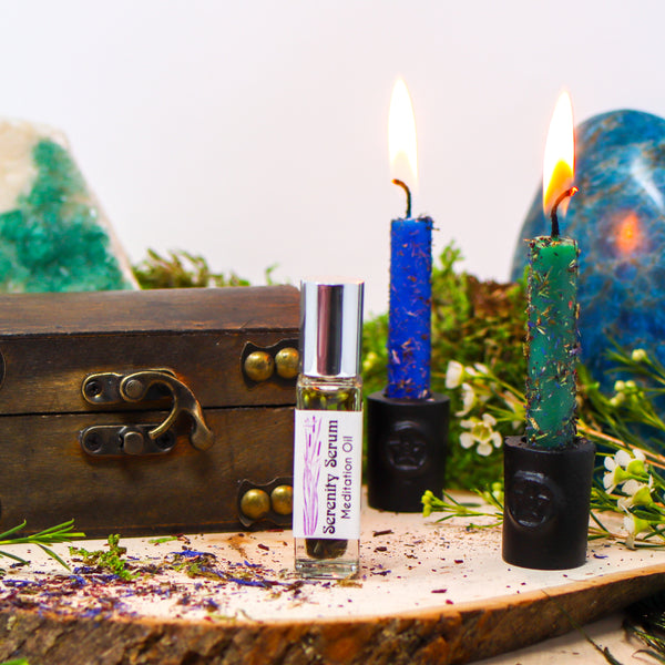 A bottle of Serenity Serum Meditation Oil between a wooden box and two burning candles dressed with herbs on a natural wood slab, with Fluorite and Blue Apatite in the background