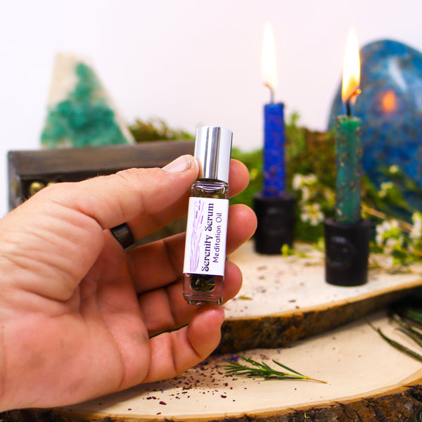 A hand holding up a bottle of Serenity Serum Meditation Oil in front of two lit candles that have been dressed with herbs, with Fluorite and Blue Apatite in the background