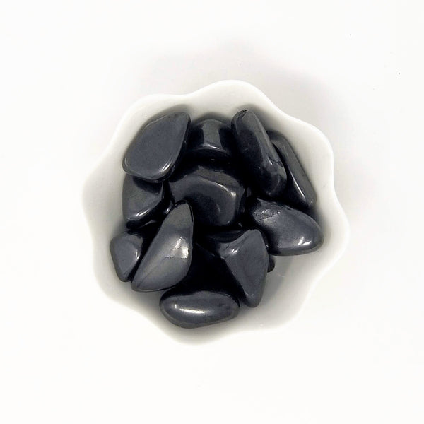 A white bowl with scalloped edges, filled to the brim with Shungite Tumbled Stones, on a white background