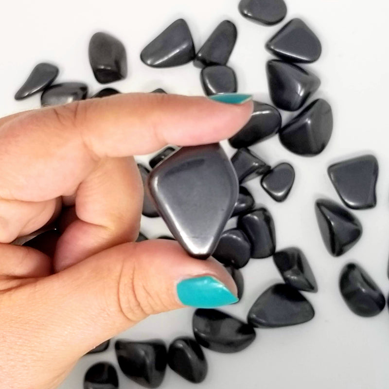 A Shungite Tumbled stone held up between a forefinger and thumb in front of more tumbled Shungite on a white background