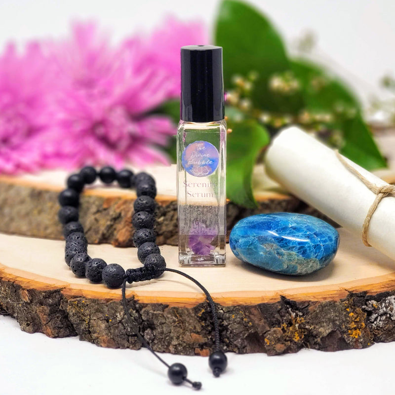 The contents of So Long Stress Morning Meditation Ritual, including Lava and Black Onyx diffuser bracelet, Serenity Serum Anointing Oil, Blue Apatite palm stone, and guided meditation scroll, displayed on two stacked natural wood slabs with fresh pink flowers in the background