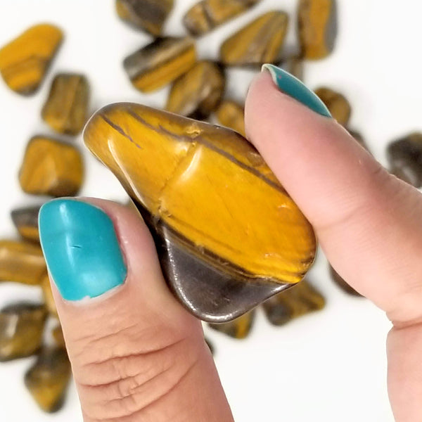 A Tiger's Eye tumbled stone held between a forefinger and thumb, with more Tiger's Eye in the background